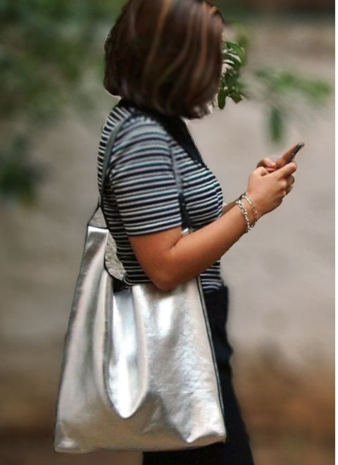 Una modelo lleva al hombro un bolso  de piel metalizada en color plata. Es un bolso grande, alegre, luminoso y con carácter.
