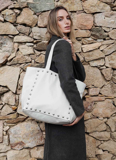 Modelo  femenina lleva al hombro nuestro bolso blanco tachuelas hecho a mano en nuestro atelier con piel de vacuno y tachas metálicas plateadas. El bolso tote tiene dos asas reforzadas de la misma piel para llevarlo al hombro o en la mano.