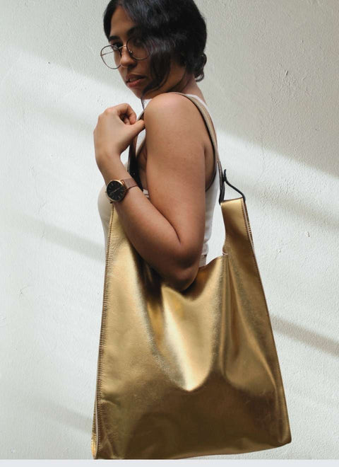 Una modelo lleva colgado al hombro un bolso metalizado confeccionado artesanalmente en nuestro taller con una preciosa piel de color oro . Es un bolso espacioso y el. que le proporciona su alegre y especial carácter. Puedes llevarlo en la mano y para mayor comodidad tiene un asa extraíble que te permite colgarlo cómodamente del hombro. Es un bolso espacioso y elegante.