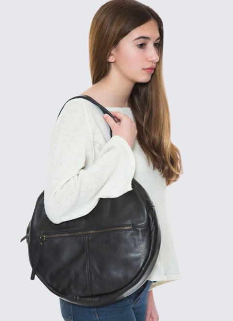 Una modelo lleva al hombro un elegante bolso redondo hecho  con cuero natural suave y liviano. El bolso tienen dos asas y un bolsillo exterior con cremallera. 